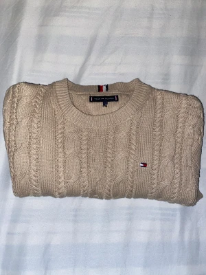 Beige stickad tröja Tommy Hilfiger - Snygg beige stickad tröja från Tommy Hilfiger med klassiskt kabelstickat mönster och rund hals. Tröjan har lång ärm och ett litet broderat Hilfiger-logo på bröstet. Perfekt för dig som gillar stilrena och tidlösa plagg med premiumkänsla. Den har en liten defekt då storlekslappen är av, men inget som syns alls. Väldigt bra skick. | säljer för 300 | Nypris - ca 1000 kr | 