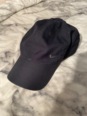Svart Nike Heritage86 keps - Säljer en stilren svart keps från Nike, modell Heritage86. Kepsen har klassisk böjd skärm och ett litet silverfärgat Nike Swoosh broderat på sidan. Tillverkad i mjuk bomull och passar de flesta tack vare justerbar storlek.