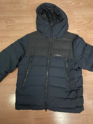 Mörkblå Sail Racing dunjacka med huva - Snygg mörkblå dunjacka från Sail Racing med huva och detaljer i grått tyg över bröstet. Jackan har två snedställda dragkedjor framtill, quiltad design och insida med GORE-TEX Infinium. Perfekt för kalla dagar och har en clean look.