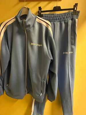 Palm Angels blå tracksuit, M - Här har vi ett blått tracksuit-set från Palm Angels med vita och svarta ränder längs sidorna, storlek M. Aldrig använd. 