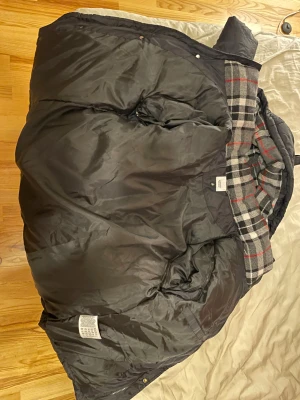 Svart pufferjacka från Burberry - Säljer en svart pufferjacka från Burberry med huva och klassiskt rutigt foder på insidan. Jackan har en patch med 'BURBERRY LONDON ENGLAND' på ärmen och dragkedja framtill. Perfekt för kalla dagar och riktigt snygg streetstil. Man kan ta bort armarna så det blir en väst, oanvänd. Skickas i original påse.
