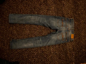 Blå jeans från Nudie Jeans - Säljet ett pat snygga Jeans från Nudie i modellen Grim Tim. Passformen är slim fit. Storlek 28:32. Det är lagning vid grenen och ett litet hål i fickan. Det får en sliten och tvättat look. Nypris runt 1600kr