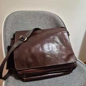 Brun portfölj från Tony Perotti - Snygg brun portfölj från Tony Perotti i äkta skinn med justerbar axelrem och flera praktiska fack. Väskan har magnetlås, dragkedja och detaljer i mässing. Insidan är fodrad med logotypmönstrat tyg och har nätficka för extra förvaring. Perfekt för laptop och dokument.