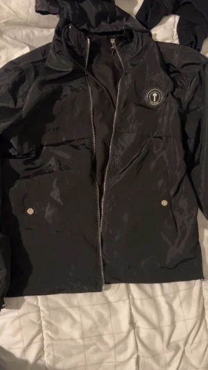 Svart vindjacka från Trapstar, storlek S - Svart vindjacka från Trapstar med stor logga broderad på ryggen och mindre logga på bröstet. Jackan har huva, dragkedja framtill, snörning och två fickor med tryckknappar. Materialet är 100% polyester säljer jackan för endast 750 vid snabba affärer 650 pågrund av har använt den ett par gånger men är fortfarande i riktigt bra skick