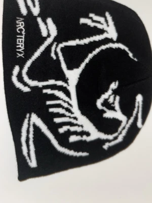 Svart mössa från Arc'teryx - Svart mössa från Arc'teryx med vit grafisk design och logga på framsidan. Mjuk och stretchig, perfekt för kalla dagar. Snygg och enkel att matcha med din vinteroutfit.