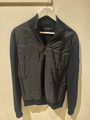 Mörkblå Cardigan J.Lindeberg - Snygg mörkblå cardigan från J.Lindeberg med quiltad framsida och stickade ärmar och rygg. Jackan har dragkedja framtill, ribbad krage och detaljerad logotyp på ärmen. Denna kommer få dig att vara tuffast på skolan! Skämt åt sidan, bara att höra av sig vid eventuella funderingar!😉