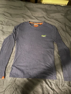 Mörkblå långärmad topp från Superdry - Säljer en mörkblå långärmad topp från Superdry Orange Label med rund hals och liten grön logotyp på bröstet. Tunn och mjuk bomullskvalitet, perfekt för chill dagar. Diskret orange detalj vid ärmslutet och snyggt tryck i nacken.