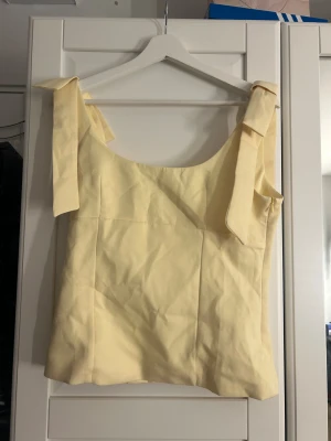 Ljusgul linnetopp med rosettband - Säljer en ljusgul linnetopp med breda axelband som knyts i rosetter. Toppen har en mjuk, rak form och är ärmlös med rundad halsringning. Materialet känns lätt och somrigt, perfekt för varma dagar. Enkel och stilren design som ger en fräsch vibe. Dragkedja på sidan och stor i storlek 