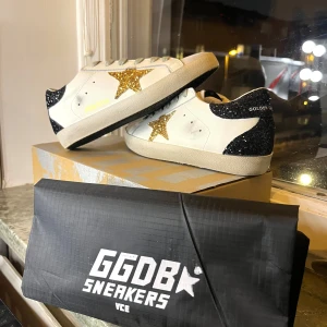 Golden Goose Superstar med guld glitter - Golden Goose sneakers i vitt skinn med grå snörning, glittrig guldstjärna på sidan och svart glitter på hälen. Platt sula och rund tå. Snygg kontrast mellan de glittriga detaljerna och den klassiska vita basen. Perfekt för dig som vill sticka ut med dina skor.