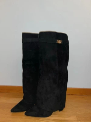 Svarta höga stövlar med spetsig tå - Säljer ett par svarta höga stövlar i mocka med spetsig tå och klack då dem ej passa mig. Snyggt band med guldfärgad detalj upptill. Perfekta för dig som vill ha en stilren och edgy look till hösten eller vintern. Bra skick, fick dom av min syster 