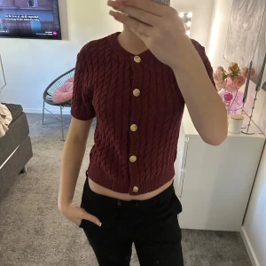 Vinröd kabelstickad kofta med guldknappar - Säljer en vinröd kabelstickad kofta med korta ärmar och snygga guldknappar framtill. Koftan är croppad och har rund halsringning. Perfekt för dig som vill ha en trendig och stilren look med lite retrovibe.