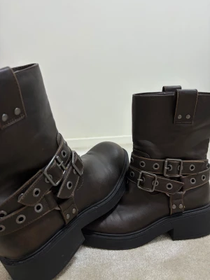 Bruna boots med dubbla spännen - Säljer ett par mörkbruna boots i skinn med rund tå och platt sula. De har två breda remmar med metallspännen och nitar runt skaftet, vilket ger en cool och robust look. Perfekt för dig som gillar lite edge i din stil. Jag har haft på mig dom minst 2 gånger sen har dom bara legat i min garderob. Helt nya typ och jättebra kvalite.
