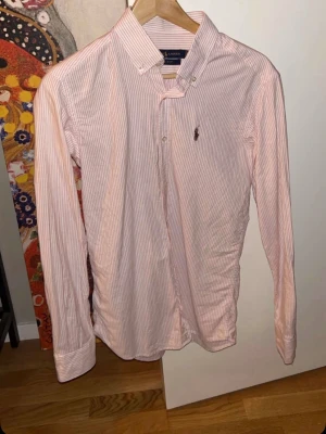 Rosa randig skjorta från Ralph Lauren - Säljer en slim fit skjorta från Ralph Lauren i storlek medium. Skjortan är ljusrosa med tunna röda ränder, har klassisk krage och knappar framtill. Broderad logga på bröstet. Perfekt för dig som gillar stilrena och snygga plagg.