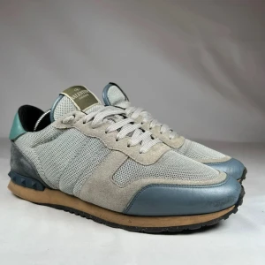 Valentino Rockrunners - Snygga Valentino Garavani Rockrunner sneakers i ljusgrå mesh och mocka med blå detaljer i skinn. Skorna har klassisk snörning, rund tå och en beige sula med svarta nitar på hälen. 