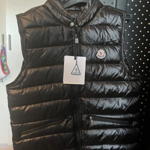 Moncler Gui Väst - Snygg svart dunväst från Moncler med glansig finish och klassisk logga på bröstet. Västen har två dragkedjefickor framtill och är helt oanvänd. Perfekt för lager-på-lager och streetstyle.