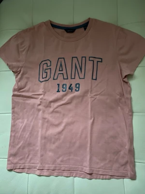 Rosa GANT t-shirt med tryck - Snygg rosa t-shirt från GANT med stort tryck 'GANT 1949' i mörkblått på bröstet. Klassisk rund hals och korta ärmar. Tillverkad i mjuk bomull som är skön mot huden. Perfekt för dig som gillar stilrena och sportiga plagg med tydlig logga.