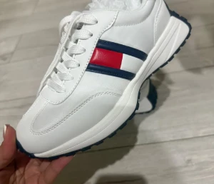 Nytt  sneakers med röd och blå detalj - Snygga vita sneakers från Tommy Hilfiger med klassisk röd, vit och marinblå detalj på sidan. Skorna har snörning, rund tå och platt sula. Perfekta för dig som gillar sportig och stilren design. Materialet är syntet och skinn.