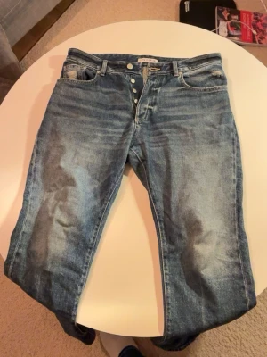 Blå jeans från Icon Denim, storlek 33 - Säljer ett par blå jeans från Icon Denim i storlek 33. Jeansen har klassisk femficksdesign, knäppning med flera knappar och en snygg tvättad look med ljusare partier. Materialet är 100% bomull och passformen är loose med raka ben. Modell: 175cm