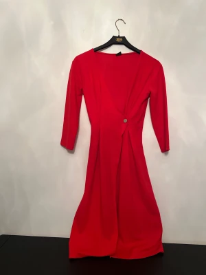 Röd omlottklänning med knapp - Snygg röd klänning med omlott-design och en silverfärgad knapp i midjan. Klänningen har trekvartsärm och v-ringad hals. Tyget är slätt och känns som polyester, vilket ger ett fint fall. Perfekt för dig som vill sticka ut med en enkel men elegant look.