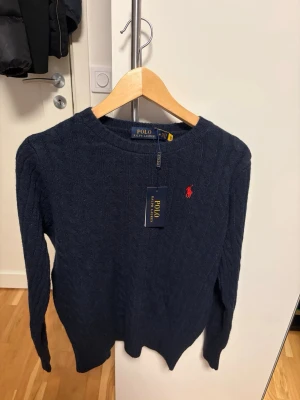 Marinblå kabelstickad tröja Polo Ralph Lauren - Säljer en marinblå kabelstickad tröja från Polo Ralph Lauren i storlek M. Tröjan har kabelstickat mönster, rund hals och lång ärm. Klassisk röd logga broderad på bröstet. Perfekt för dig som gillar stilrena och tidlösa plagg. Storlek M, aldrig använd, prislappen är kvar. Pris kan diskuteras!