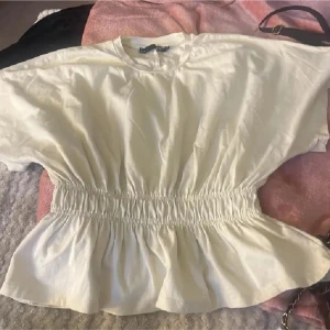 Vit blus med smock i midjan - Säljer en vit blus med rund hals och korta ärmar. Toppen har smockad midja som ger en snygg volang-effekt nedtill. Perfekt för dig som gillar en feminin och chill stil. Materialet känns mjukt och lätt, troligen bomull.