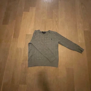 Grå stickad tröja från Polo Ralph Lauren - Snygg grå stickad tröja från Polo Ralph Lauren med klassisk kabelstickad design och rund hals. Tröjan har långärmade ärmar och en liten broderad logga på bröstet. Perfekt för dig som gillar stilrena och tidlösa plagg.