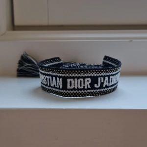 Christian Dior J'Adior armband - Snyggt armband från Christian Dior med texten 'J'Adior' broderad i vitt. Armbandet har en vävd design med vita detaljer och avslutas med två coola tofsar i blått och vitt. Perfekt accessoar för att lyfta din outfit med lite lyxig vibe. Justerbar storlek genom att dra i snörena (Passar alla) ✌️ Hör av dig vid minsta fråga!