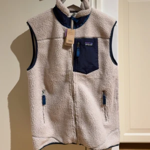 Patagonia Classic Retro-X fleeceväst M - Säljer en  beige och marinblå Patagonia Classic Retro-X fleeceväst i storlek M. Säljer då den var för liten på mig så den har aldrig varit använd förutom när jag provade den hemma. Västen har mjuk teddyfleece, hög krage, hel dragkedja och tre fickor med blå detaljer. Perfekt för lager-på-lager.