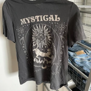 Mystical svart t-shirt med print - Svart t-shirt med coolt vitt print framtill med sol, måne, stjärnor och leopard samt texten 'MYSTICAL'. Rund hals och korta ärmar. Materialet känns som mjuk bomull, perfekt för dig som gillar unika och mystiska motiv.