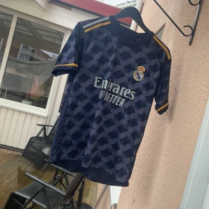 Real Madrid Bellingham bortatröja - Snygg Real Madrid bortatröja med Bellingham 5 på ryggen. Tröjan är mörkblå med unikt mönster, gula detaljer på ärmar och axlar samt klubbmärke och sponsortryck framtill. Tillverkad i lätt och ventilerande polyester, perfekt för fotboll eller supporterliv.