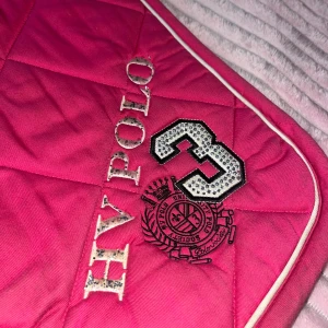 Hv Polo schabrak - Bilderna visar en rosa HV Polo schabrak med broderad logotyp och siffran 3 i glittrande detaljer. Schabraket har en quiltad design och vita kanter. Viss användning syns, men överlag i gott skick. Perfekt för dig som vill ha en snygg och sportig look till din häst.