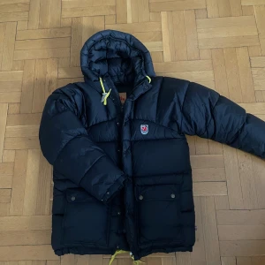 Svart dunjacka från Fjällräven - Svart pufferjacka från Fjällräven med huva och gula snören. Jackan har två stora fickor framtill med knappar och klassisk Fjällräven-logga på bröstet. Fylld med dun och fjäder, perfekt för kalla dagar. 