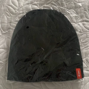 Svart mössa från E.ON - Svart, enkel och stilren mössa med en röd etikett där det står 'e.on' på kanten. Mössan har en klassisk form och är tillverkad i mjukt material som passar perfekt för kyliga dagar. Snygg accessoar för dig som gillar minimalistisk stil.