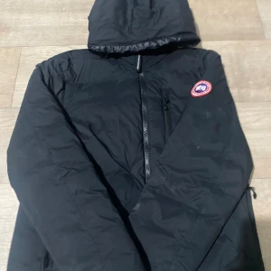 Svart jacka från Canada Goose - Svart jacka från Canada Goose med luva och klassisk logga på ärmen. Jackan har quiltad insida, dragkedja framtill och en bröstficka med dragkedja. Perfekt för kalla dagar och har en clean, stilren look som funkar till streetwear. Hör gärna av dig för lflera frågor. Ny pris 9000kr.