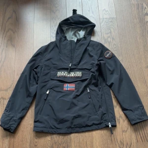 Svart vindjacka från Napapijri - Svart vindjacka från Napapijri med huva, halv dragkedja och stor logga på bröstet samt norsk flagga. Jackan har två dragkedjefickor framtill och patch på ärmen. Halv zip. Perfekt för blåsiga dagar och streetwear-stil. Skickar oftast inom 24h📦