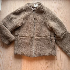 Beige teddyjacka med dragkedja - Mysig beige teddyjacka med hel dragkedja framtill och långärmad design. Jackan har en fluffig och mjuk yta i teddy-material och raka sömmar som ger en clean look. Perfekt för kyliga dagar och enkel att matcha med olika outfits.