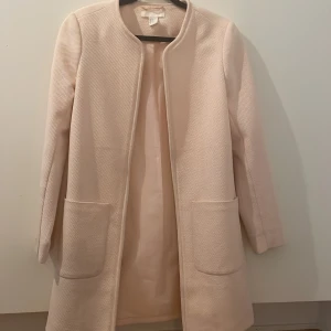 Ljusrosa kappa från H&M, stl 34 - Stilren ljusrosa kappa från H&M i storlek 34. Jackan har rak passform, stora fickor framtill och är tillverkad i strukturerad polyesterblandning. Fodrad insida och klassisk, öppen design utan knäppning. Perfekt för dig som gillar minimalistisk och clean stil.