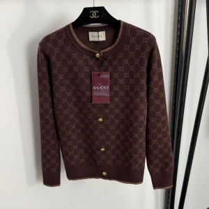 Brun kofta från Gucci med logomönster - Säljer en brun kofta från Gucci med ikoniskt GG-logomönster över hela tröjan. Koftan har rund halsringning, guldiga knappar framtill och ribbade muddar. Perfekt för dig som vill ha en lyxig och trendig look.