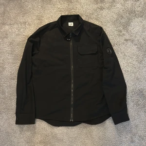 Svart overshirt från C.P. Company - Svart overshirt från C.P. Company i storlek M. Snygg design med zip framtill, klassisk krage och en ficka på bröstet. Ikonisk linsdetalj på ärmen. Materialet känns lätt och smidigt, perfekt för lager-på-lager. Passformen är dock lite liten så jag skulle nog säga att den passar S / XS. Den är i mycket fint skick men har några små fläckar som syns på bilderna. Hör av er om ni undrar något. Priset går att diskutera. 