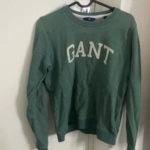 Grön sweatshirt från GANT - Snygg grön sweatshirt från GANT med klassisk rund halsringning och stora vita bokstäver framtill. Tröjan har långa ärmar och ribbade muddar vid ärmslut och nederkant. Perfekt för en chill och stilren look.