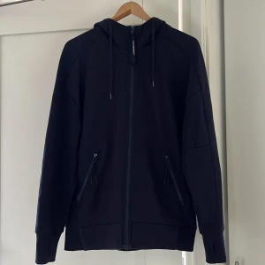 Mörk blå hoodie med goggles från C.P. Company - Mörk blå hoodie från C.P. Company med ikoniska goggles i huvan. Tröjan har hel dragkedja framtill, två fickor med dragkedja och snörning i huvan. Tillverkad i mjuk bomull och har en normal passform med långa ärmar. Perfekt för dig som gillar streetwear och unika detaljer. Pris kan diskuteras 