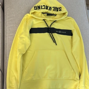 Gul hoodie från Sail Racing - Gul hoodie från Sail Racing med svart logga på huvan och ett svart band över bröstet. Tröjan har snörning i huvan, stor magficka och långa ärmar. Perfekt för dig som gillar sportig och färgstark stil.
