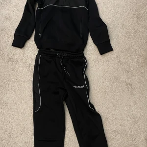Svart set från McKenzie - Svart set från McKenzie med vita detaljer längs benen och logga på låret. Byxorna har snörning i midjan och är gjorda i mjukt material som passar perfekt för chill eller träning. Snygg och enkel design med bekväm passform.