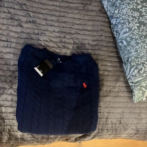 Marinblå stickad tröja Polo Ralph Lauren - Snygg marinblå stickad tröja från Polo Ralph Lauren med klassisk kabelstickning och röd broderad logga på bröstet. Rund hals och ribbade muddar vid ärmslut och nederkant. Perfekt för dig som gillar stilrena och tidlösa plagg. Hör av er gällande frågor