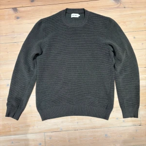 Mörkgrå stickad tröja East West - Snygg mörkgrå stickad tröja från East West med diskret ribbad struktur. Rund halsringning och långa ärmar ger en clean och stilren look. Perfekt för dig som gillar enkel design med lite edge.