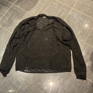 Svart leopardmönstrad skjorta ICHI - Svart transparent skjorta från ICHI i storlek M med coolt leopardmönster. Skjortan har lång ärm och en loose passform, perfekt för dig som gillar att sticka ut. Materialet känns lätt och luftigt, och den har en snygg v-ringad hals. Riktigt trendig vibe!