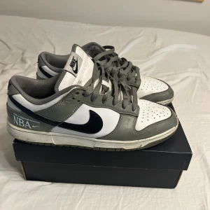 Nike Dunk Low NBA grå/vita sneakers - Nike Dunk Low sneakers med NBA-detalj, i grått och vitt med svart swoosh. Klassisk låg modell med snörning och perforerad tå. Ovandel i skinn och textil, gråa skosnören och vit mellansula. Perfekta för dig som gillar streetwear och sportig stil.