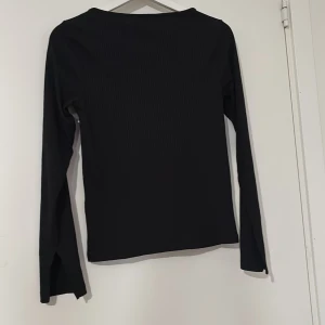 Svart långärmad topp - En stilren svart långärmad topp med diskret ribbad struktur. Modellen har en rak passform och är tillverkad i ett mjukt syntetmaterial som känns skönt mot huden. Perfekt att matcha med jeans eller kjol för en clean look.
