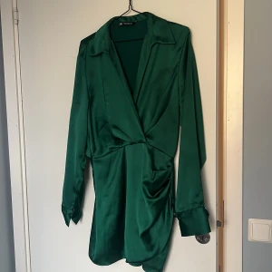 Grön omlottklänning från Zara - Snygg grön kortklänning från Zara i glansigt satinmaterial. Klänningen har omlott-design, markerad midja och långa ärmar med breda manschetter. Krage och lätt drapering framtill ger en cool och modern vibe. Perfekt för dig som gillar att sticka ut med färg.
