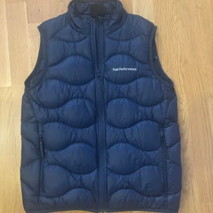 Mörkblå dunväst Peak Performance - Snygg mörkblå dunväst från Peak Performance med quiltad design och hög krage. Västen har två dragkedjefickor framtill och stängs med dragkedja. Perfekt för lager-på-lager och ger en sportig vibe.
